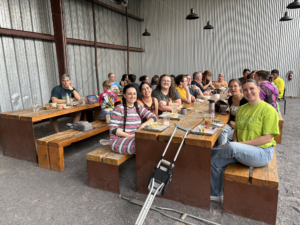 Photo of a portion of the group from the sawmill social sitting around long wooden picnic tables. Several are smiling at the camera.